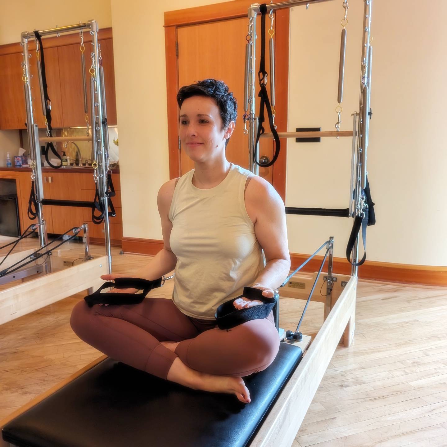 A woman sits on the reformer preparing to do the reaching exercise.